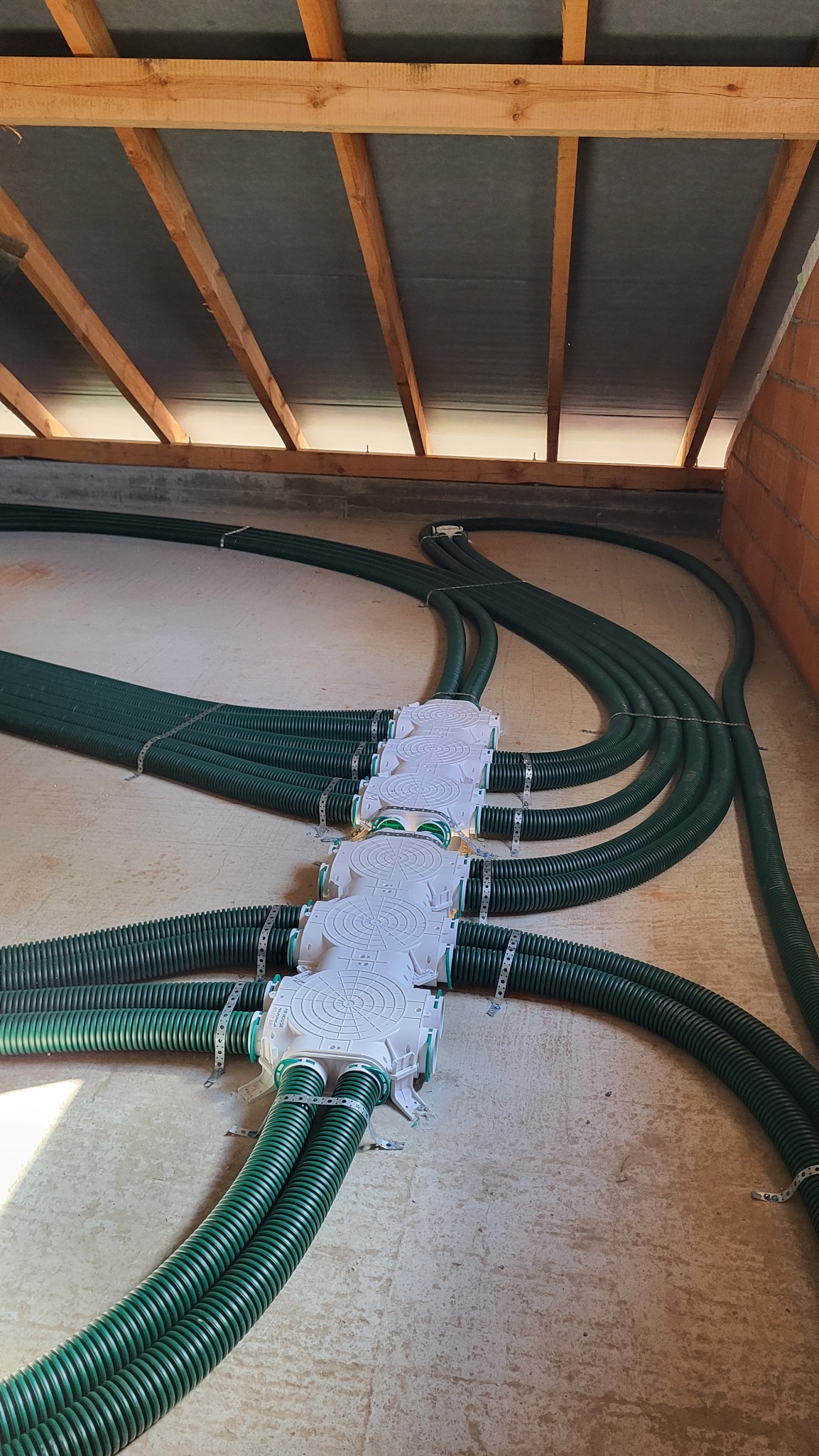 Rozprowadzenie zielonych, karbowanych przewodów wentylacyjnych pod stropem, połączonych białymi rozdzielaczami, widoczne metalowe obejmy mocujące.