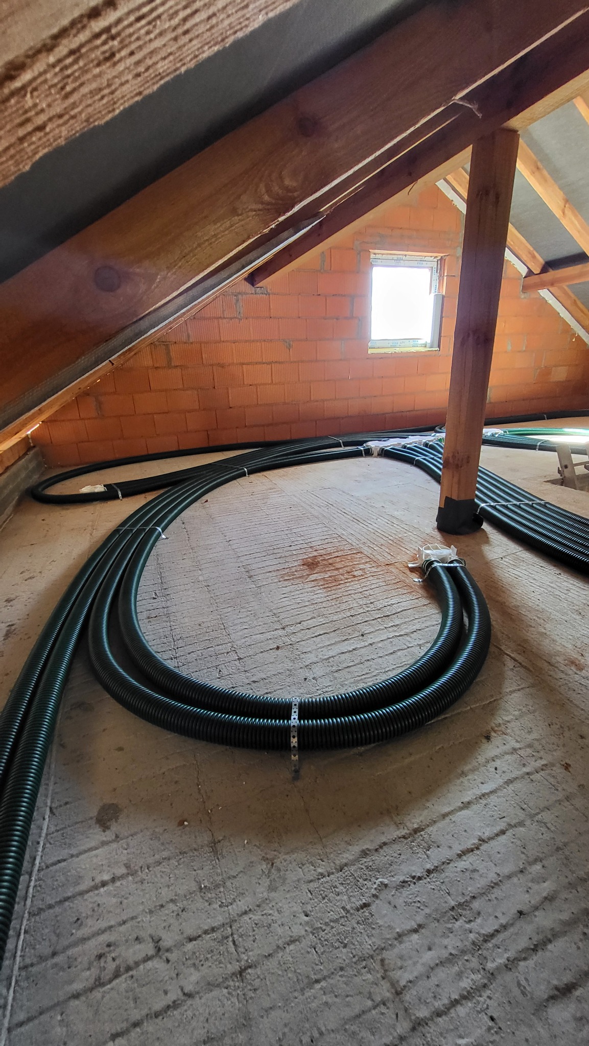 Układanie czarnych, karbowanych rur wentylacyjnych w surowym stanie poddasza z widoczną ceglaną ścianą i oknem dachowym