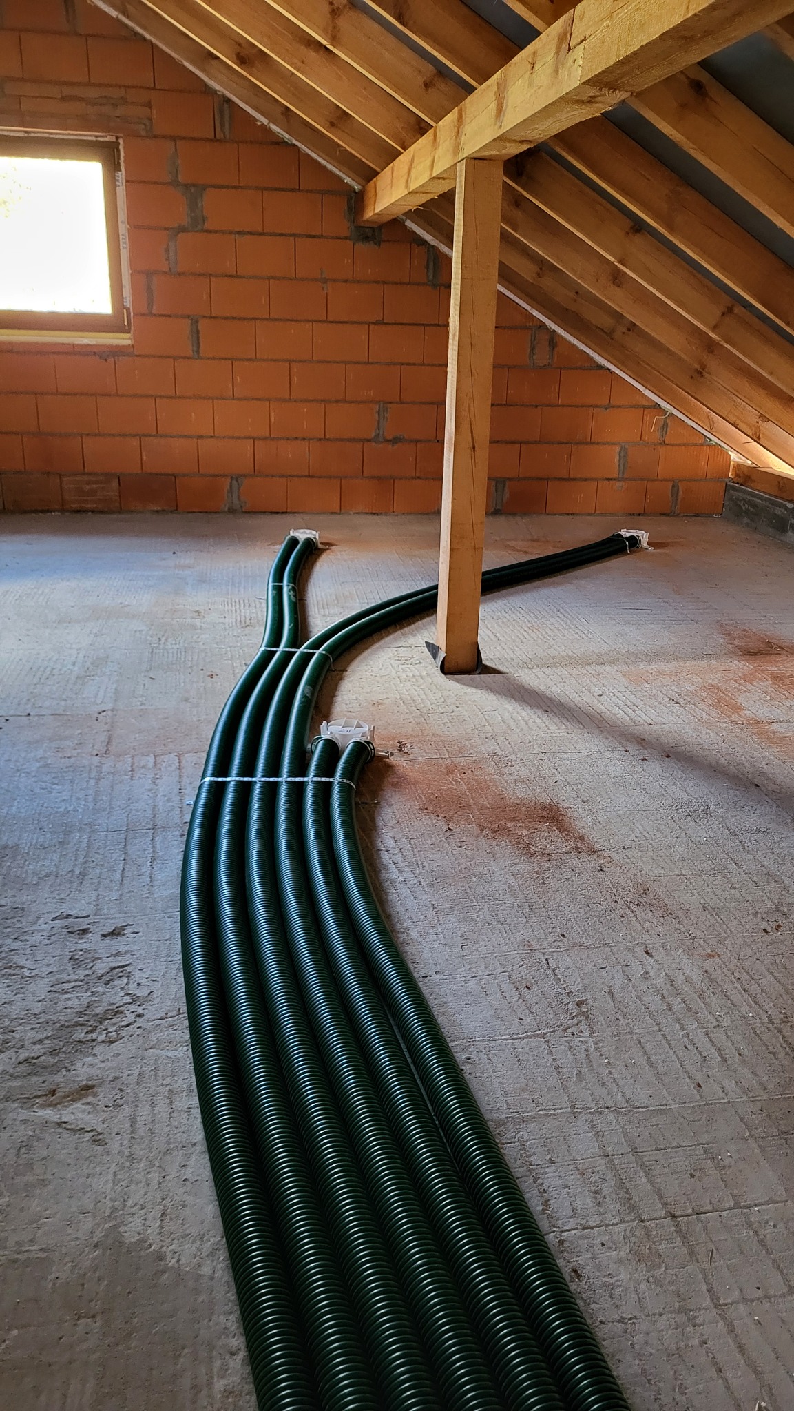 Zielone, karbowane rury wentylacyjne prowadzone po podłodze poddasza w surowym stanie, widoczne cegły, drewniana konstrukcja dachu i małe okno.