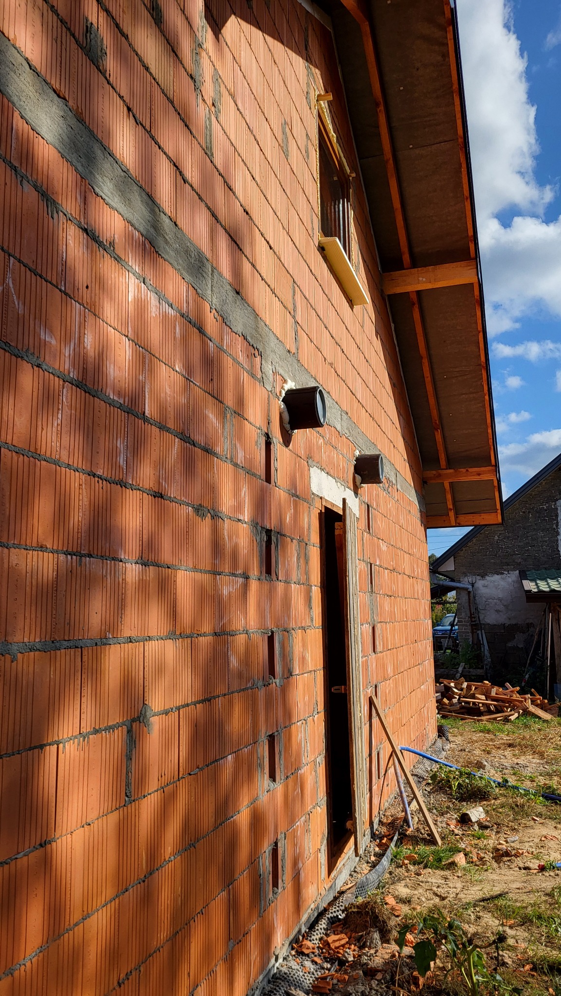 Surowa ściana budynku z czerwonej cegły z widocznymi dwoma okrągłymi wlotami wentylacyjnymi, okno z drewnianą ramą i fragment dachu w trakcie budowy, na zewnątrz widoczne ślady po budowie.