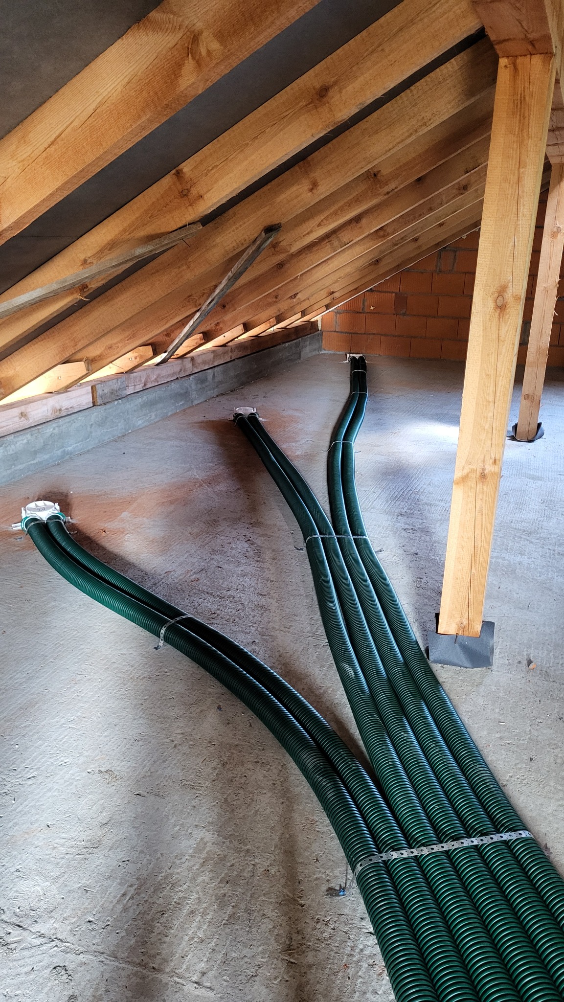 Zielone, karbowane rury wentylacyjne ułożone na betonowej podłodze poddasza z drewnianą konstrukcją dachu i fragmentem murowanej ściany w tle.