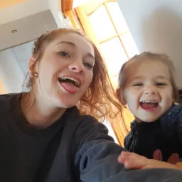 Kobieta z rudymi włosami i mała dziewczynka uśmiechają się do kamery, siedząc przy drewnianym stole w jasnym pokoju.