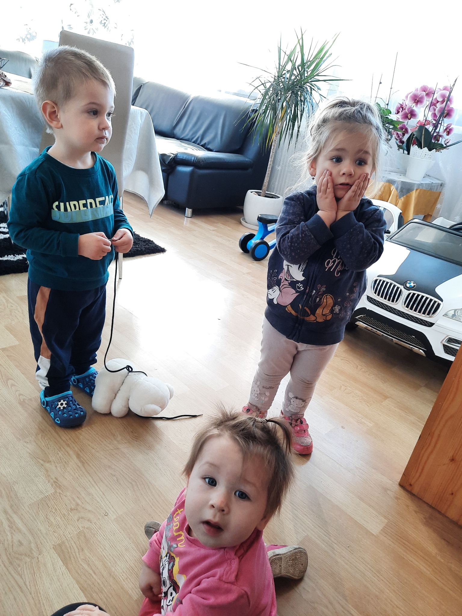 Troje dzieci bawi się w jasnym pokoju w Poznaniu: chłopiec ciągnie pluszowego misia na sznurku, dziewczynka stoi z rękami na policzkach, a najmłodsze dziecko siedzi na podłodze i patrzy w obiektyw.