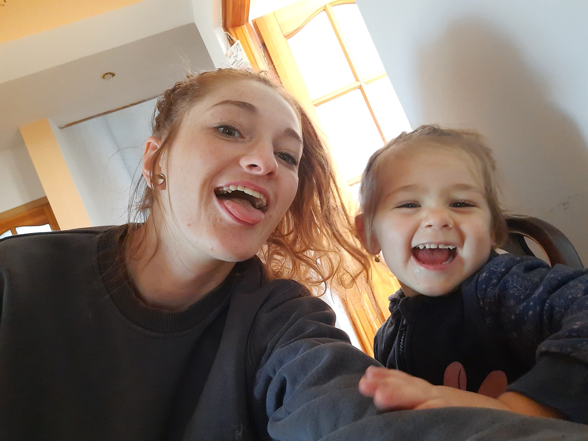 Kobieta z rudymi włosami i mała dziewczynka uśmiechają się do kamery, siedząc przy drewnianym stole w jasnym pokoju.