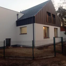 Nowoczesny dom jednorodzinny z dachem dwuspadowym, pokrytym blachą w kolorze grafitowym i brązowym, z elementami obróbek blacharskich i minimalistyczną elewacją w kolorze białym, ogrodzony siatką.