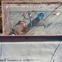 Pracownik w kombinezonie, na dachu pokrytym papą, używa palnika do zgrzewania, widoczna butla gazowa i narzędzia, ujęcie z góry.