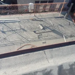 Fragment dachu płaskiego w trakcie naprawy, widoczne narzędzia, częściowo usunięta papa i przygotowane elementy do montażu, cień ozdobnej balustrady.