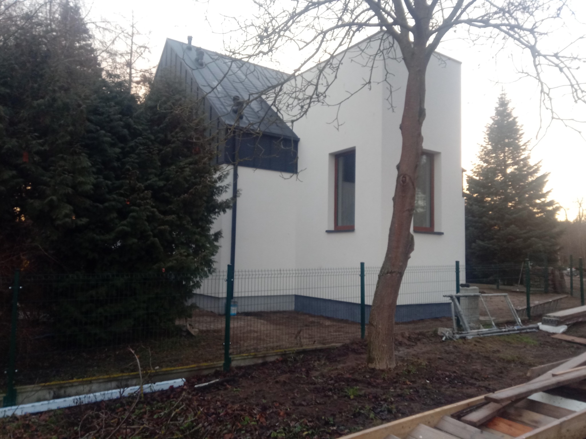 Nowoczesny dom z dwuspadowym, ciemnym dachem blachodachówkowym, białą elewacją i brązowymi oknami, otoczony zielonymi drzewami i ogrodzeniem panelowym, sfotografowany w pochmurny dzień.