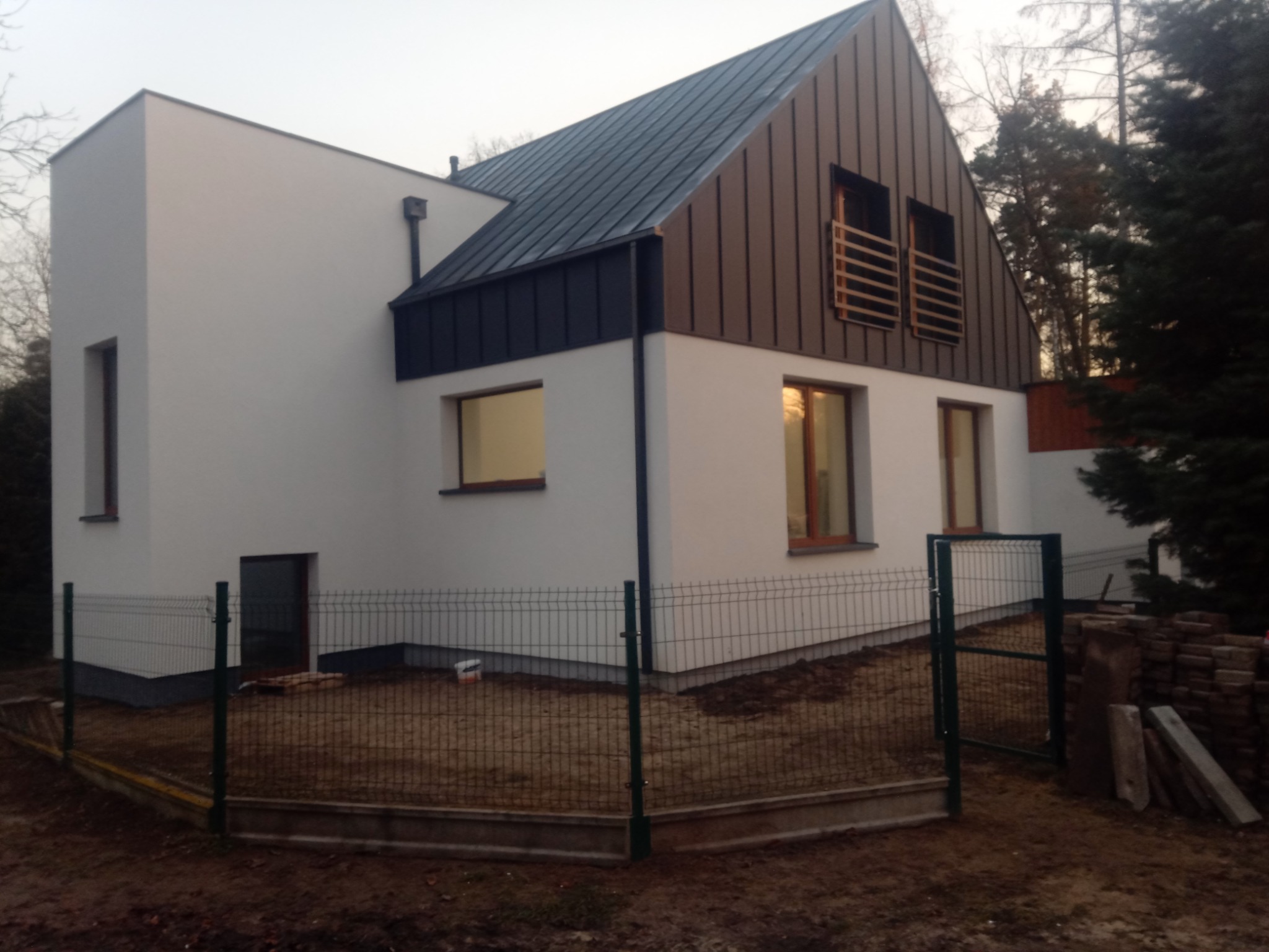 Nowoczesny dom jednorodzinny z dachem dwuspadowym, pokrytym blachą w kolorze grafitowym i brązowym, z elementami obróbek blacharskich i minimalistyczną elewacją w kolorze białym, ogrodzony siatką.