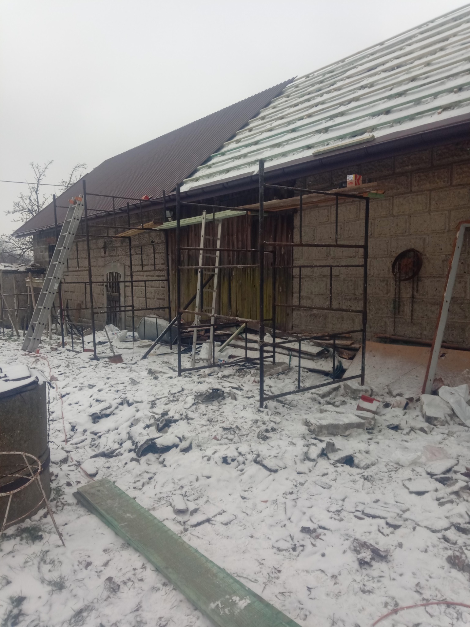 Częściowo zdjęty dach z brązową blachodachówką, widoczna więźba i rusztowanie, teren wokół budynku pokryty śniegiem i resztkami materiałów budowlanych.