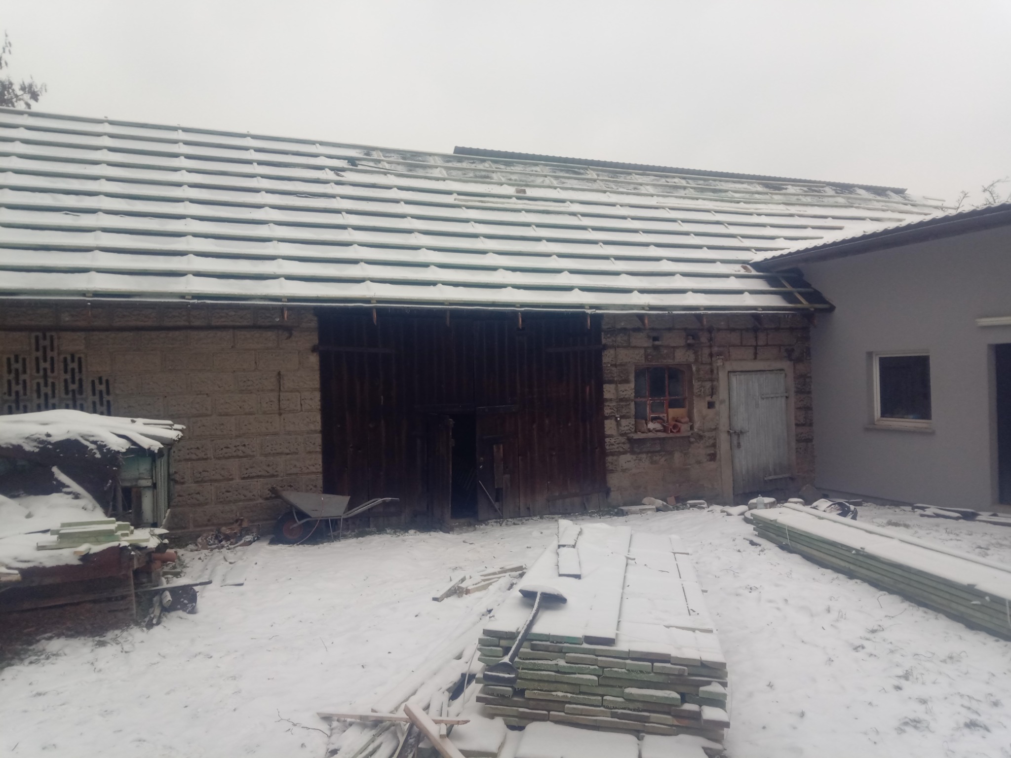 Częściowo ośnieżony, szary dach z blachy trapezowej na murowanym budynku gospodarczym z drewnianymi wrotami, obok sterta desek i narzędzia w zimowej scenerii.