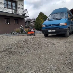 Przygotowanie podłoża pod kostkę brukową: ubijarka wibrująca obok pryzmy żwiru na tle domu z balkonami i niebieskiego busa VW.