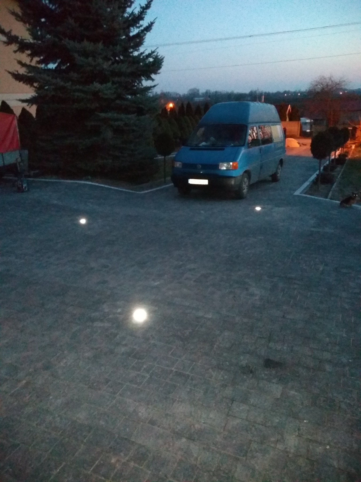 Niebieski van zaparkowany na posesji z kostki brukowej oświetlonej punktowymi lampami, widoczny świerk i inne drzewa w tle, zmierzch.