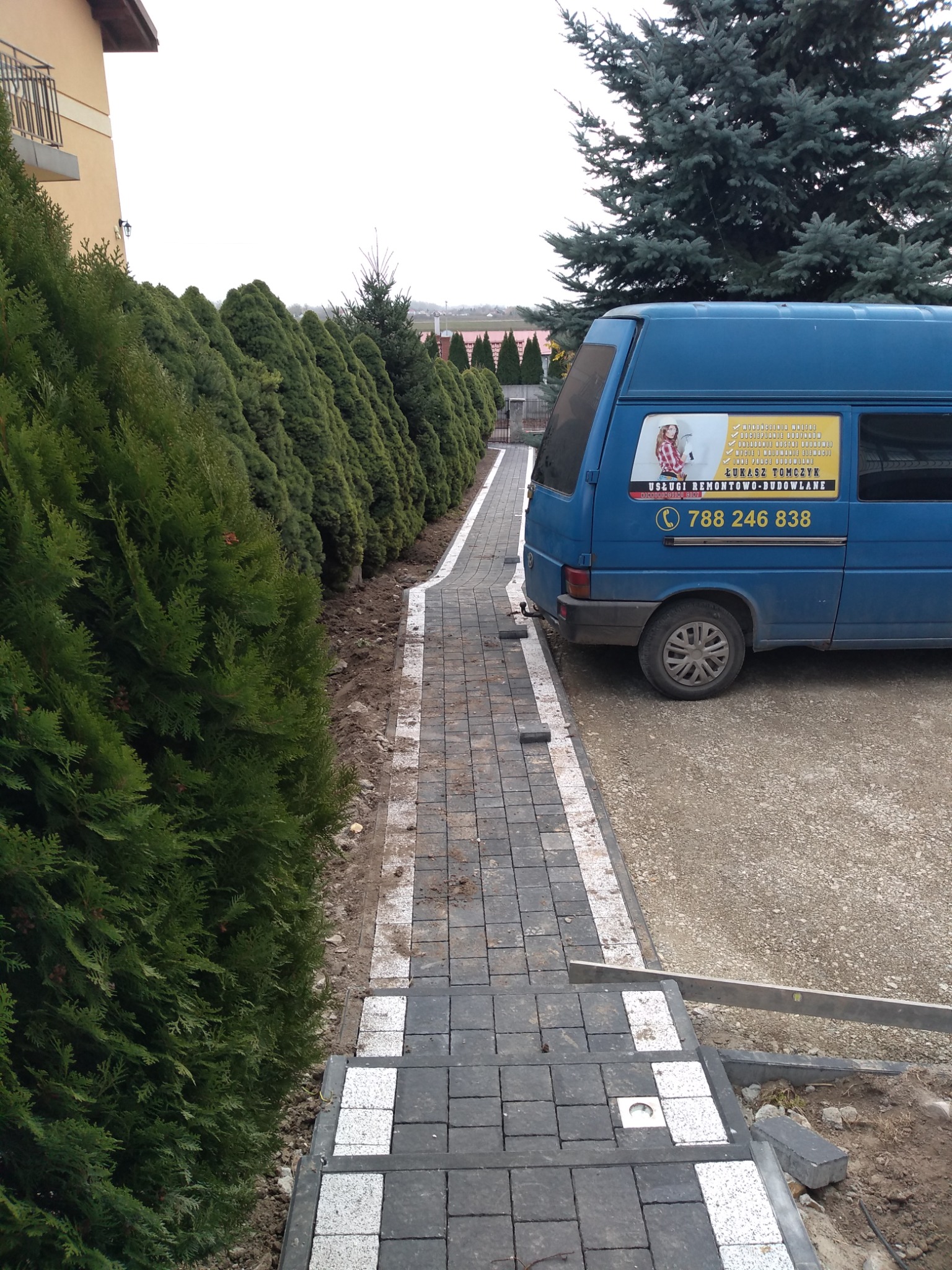 Układanie kostki brukowej w Tarnobrzegu, widok na nowo wyłożoną ścieżkę z szarej kostki z białymi akcentami, obok rząd tuj i firmowy bus z reklamą usług remontowo-budowlanych.