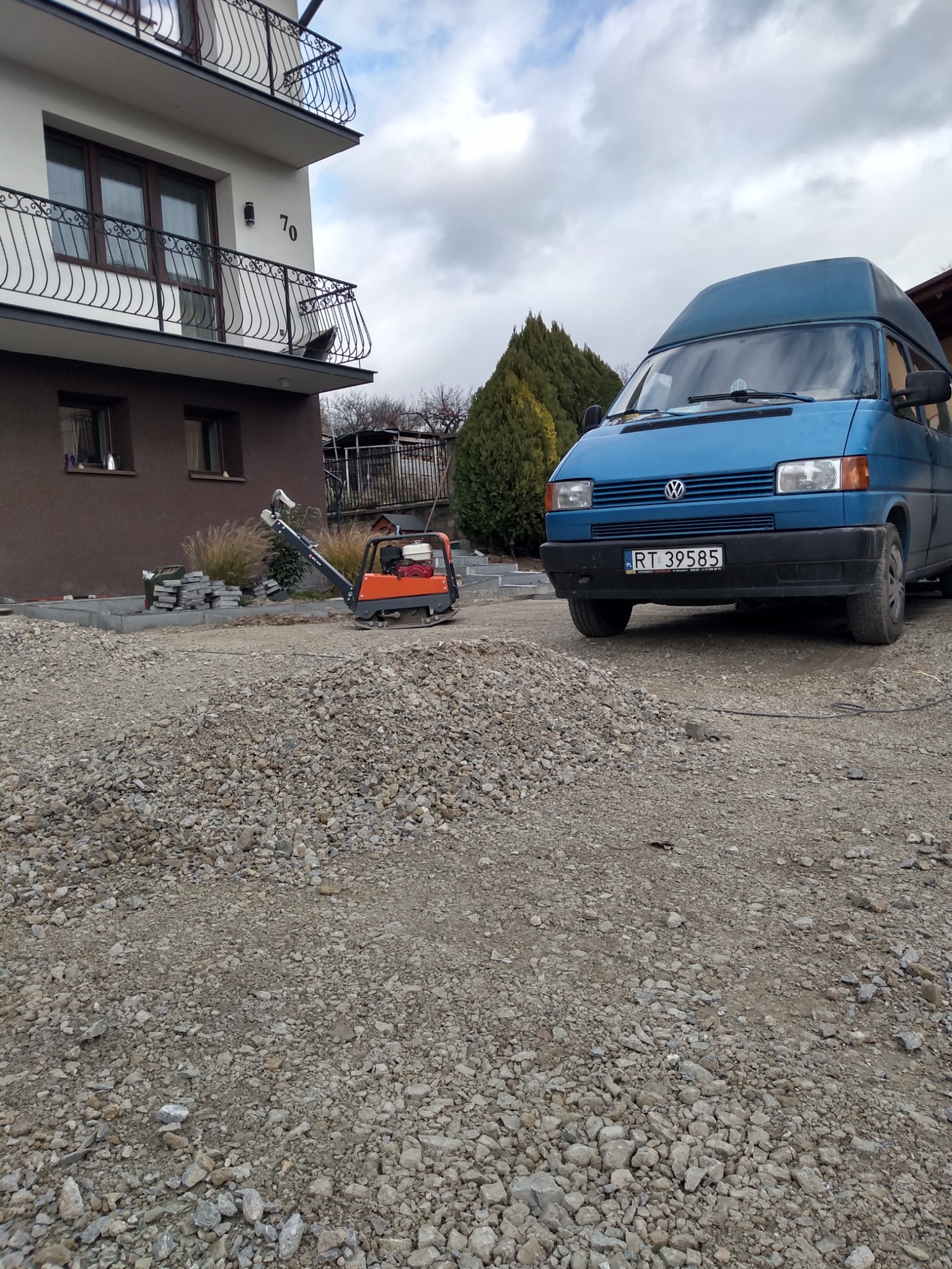 Przygotowanie podłoża pod kostkę brukową: ubijarka wibrująca obok pryzmy żwiru na tle domu z balkonami i niebieskiego busa VW.