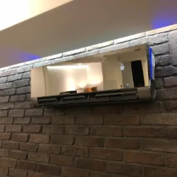 Nowoczesny klimatyzator z lustrzanym panelem frontowym zamontowany na ścianie z cegły w stylu loftowym. Na panelu widoczny wyświetlacz z cyfrą 18.