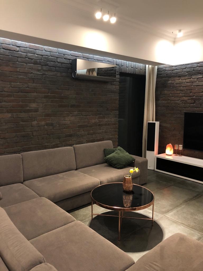 Nowoczesny klimatyzator kasetonowy zamontowany nad sofą w salonie z ceglaną ścianą, oświetleniem punktowym i okrągłym stolikiem kawowym.
