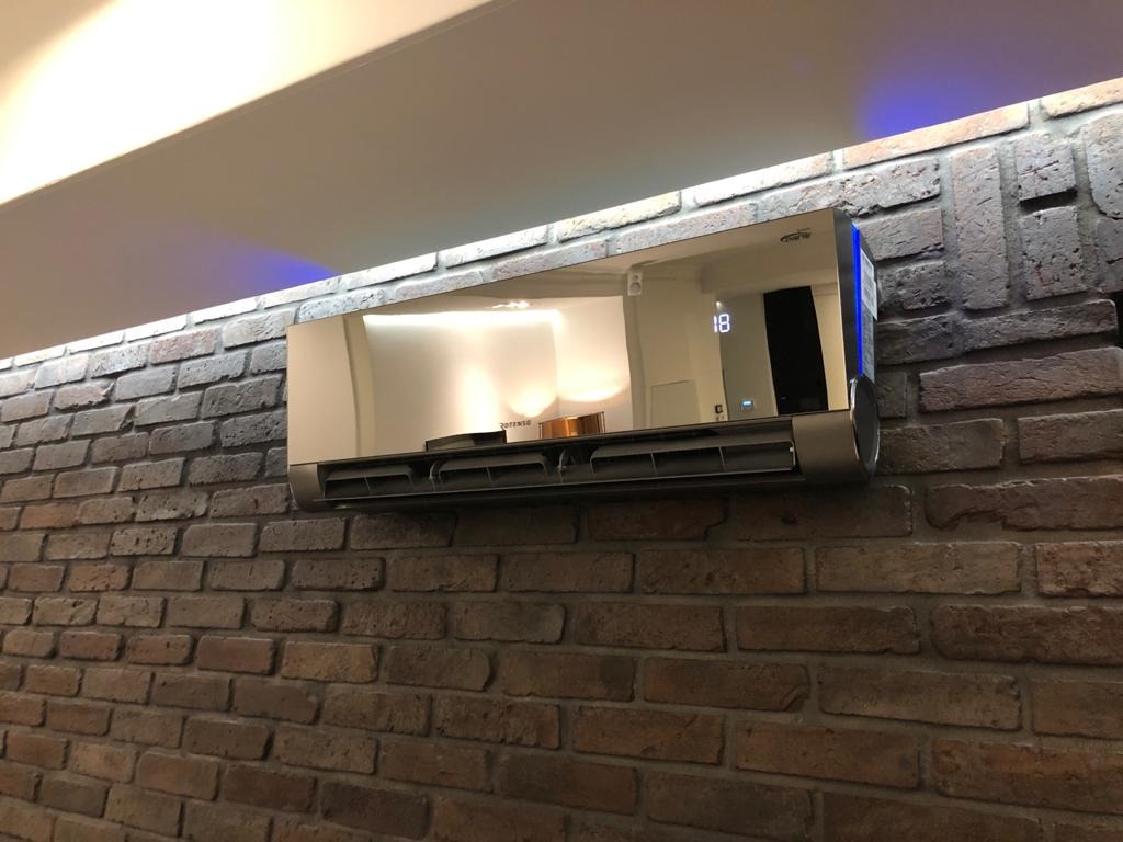 Nowoczesny klimatyzator z lustrzanym panelem frontowym zamontowany na ścianie z cegły w stylu loftowym. Na panelu widoczny wyświetlacz z cyfrą 18.