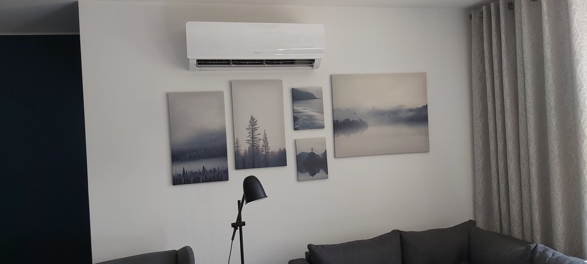 Biały klimatyzator ścienny w salonie z minimalistycznym wystrojem, wiszący nad sześcioma obrazami z motywami leśnymi i jeziornymi w szarościach, oświetlony czarną lampą stojącą.