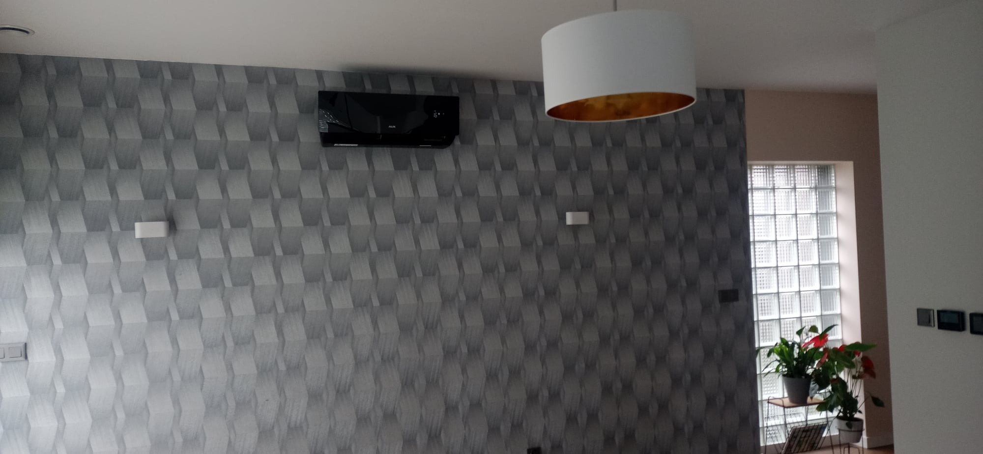 Czarny klimatyzator ścienny zamontowany na ścianie z trójwymiarową tapetą, widoczna lampa sufitowa i okno z luksferów z roślinami.