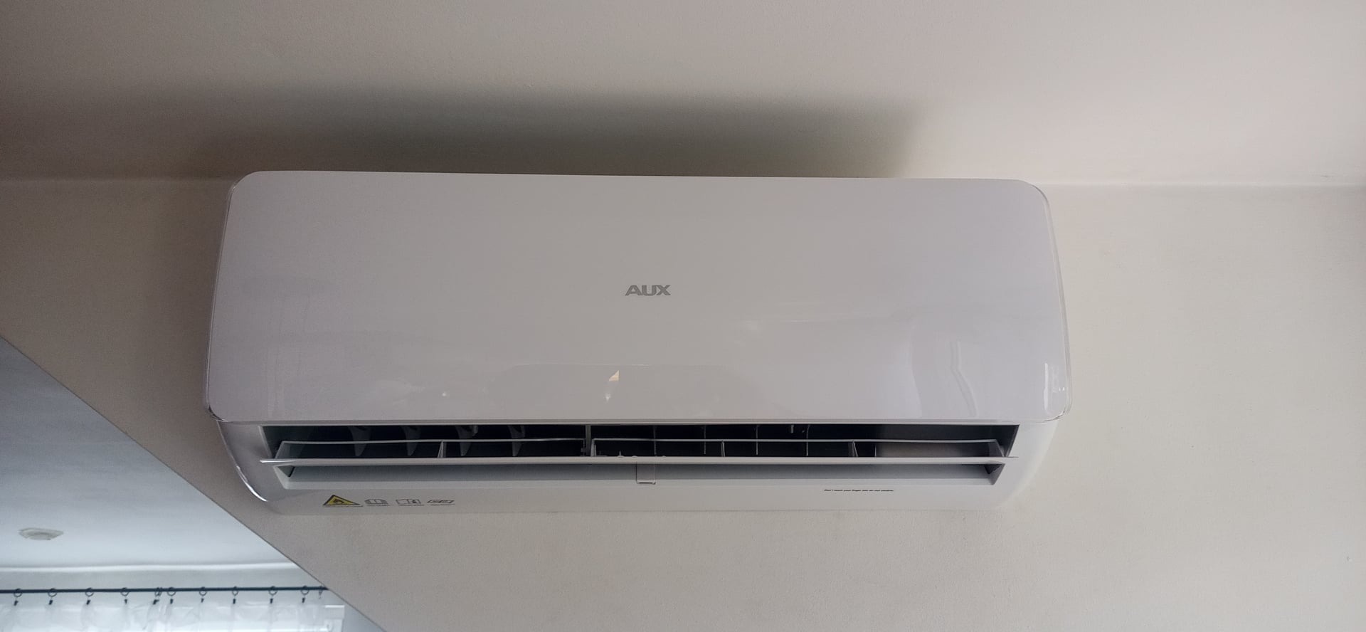 Biała klimatyzacja AUX zamontowana na jasnej ścianie w pomieszczeniu. Widoczne żaluzje i logo producenta.