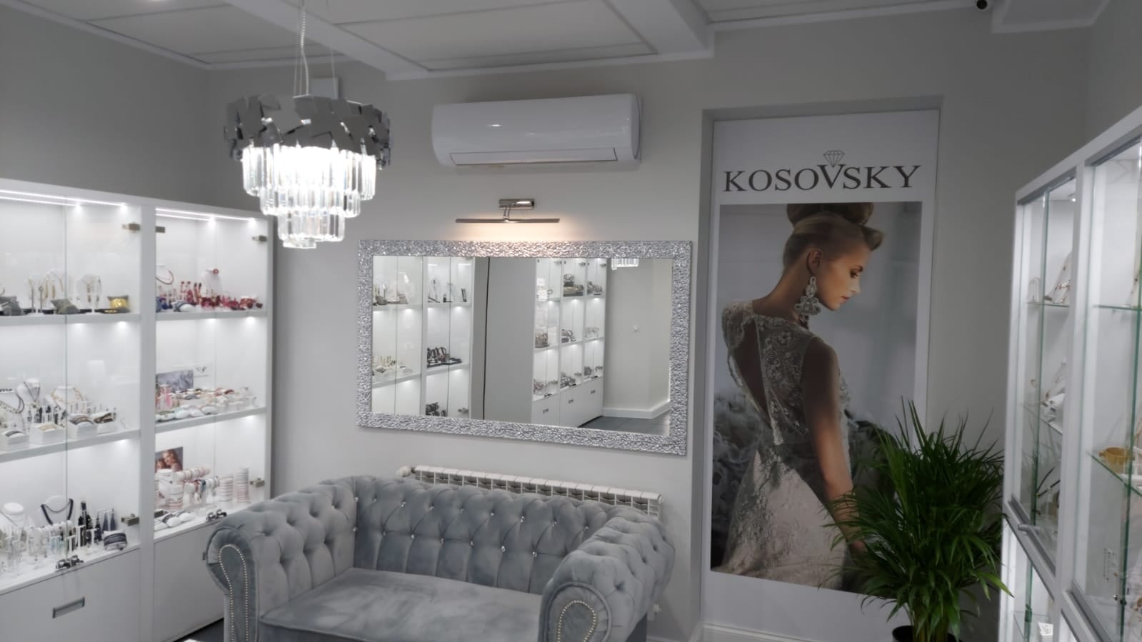 Wnętrze butiku jubilerskiego z klimatyzatorem ściennym, szarym welurowym siedziskiem, lustrem w ozdobnej ramie i oświetlonymi gablotami z biżuterią, obok plakat reklamowy.