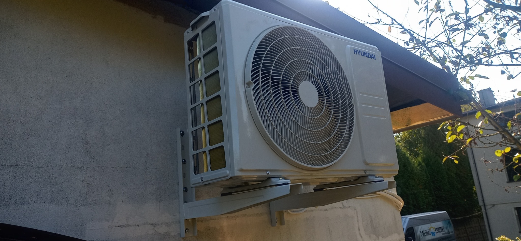 Zewnętrzna jednostka klimatyzacji Hyundai zamontowana na ścianie budynku, widoczne elementy montażowe oraz fragment dachu i drzewa w tle.