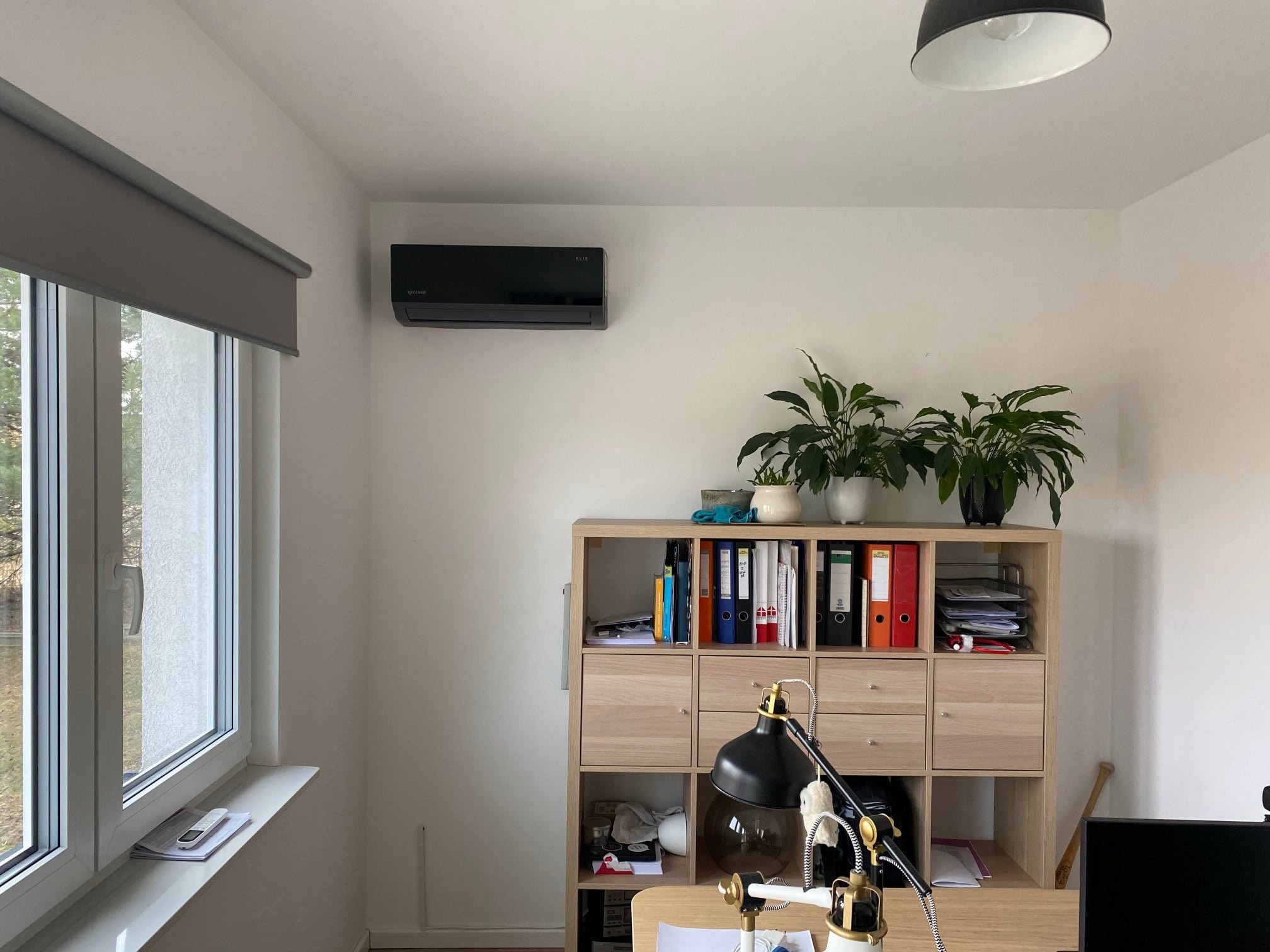Nowoczesny, czarny klimatyzator zamontowany na białej ścianie w jasnym pokoju biurowym, widoczne biurko, regał z segregatorami, roślina doniczkowa i okno z żaluzjami.