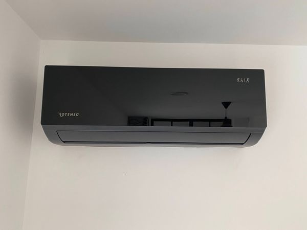 Czarny klimatyzator Rotenso Elis zamontowany na białej ścianie, widoczne odbicie lampy w obudowie.