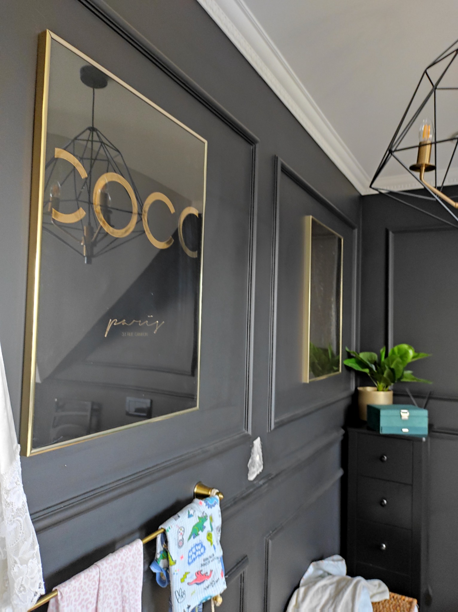 Ściana w ciemnoszarym kolorze z dekoracyjnymi listwami, ozdobiona złotymi ramami z grafikami i lustrem. Na wieszaku ręczniki w pastelowych kolorach. W rogu pokoju czarna komoda z rośliną doniczkową...