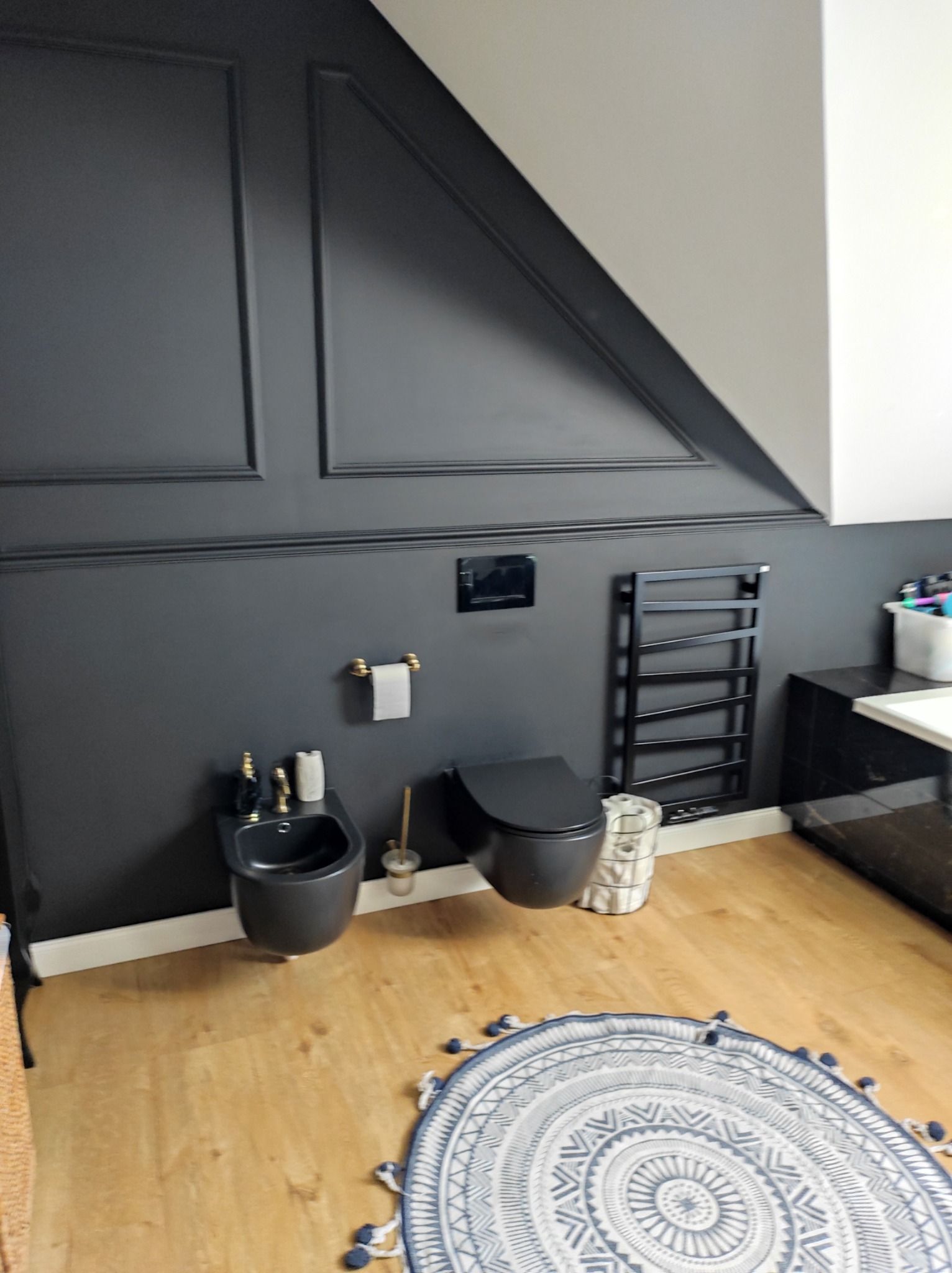 Elegancka łazienka w odcieniach szarości z drewnianą podłogą, bidet i toaleta w kolorze grafitowym, okrągły dywan z frędzlami, dekoracyjne listwy ścienne.