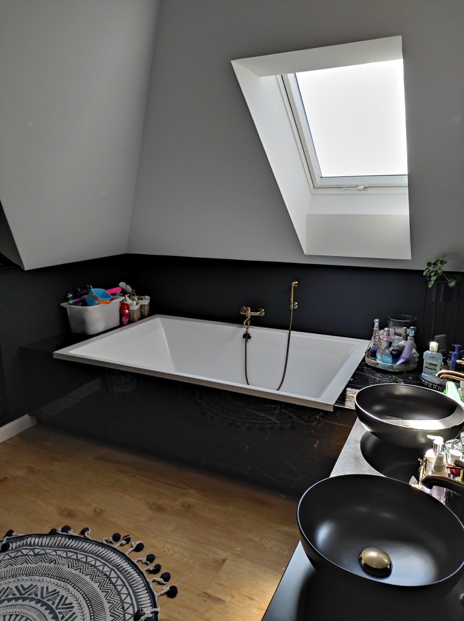 Nowoczesna łazienka na poddaszu z czarnymi płytkami imitującymi marmur, białą wanną pod oknem dachowym i dwiema czarnymi umywalkami nablatowymi.