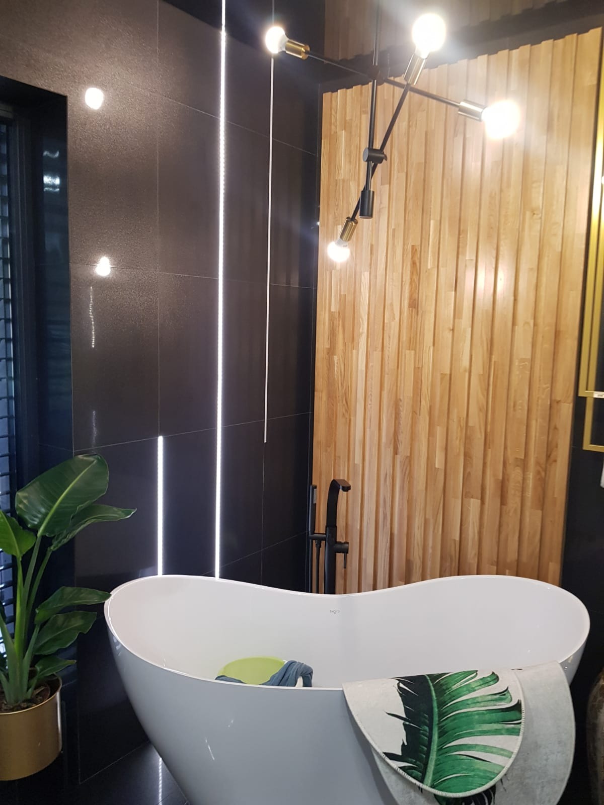 Nowoczesna łazienka z wolnostojącą wanną, czarnymi płytkami z podświetleniem LED, drewnianą ścianą dekoracyjną i designerską lampą sufitową.