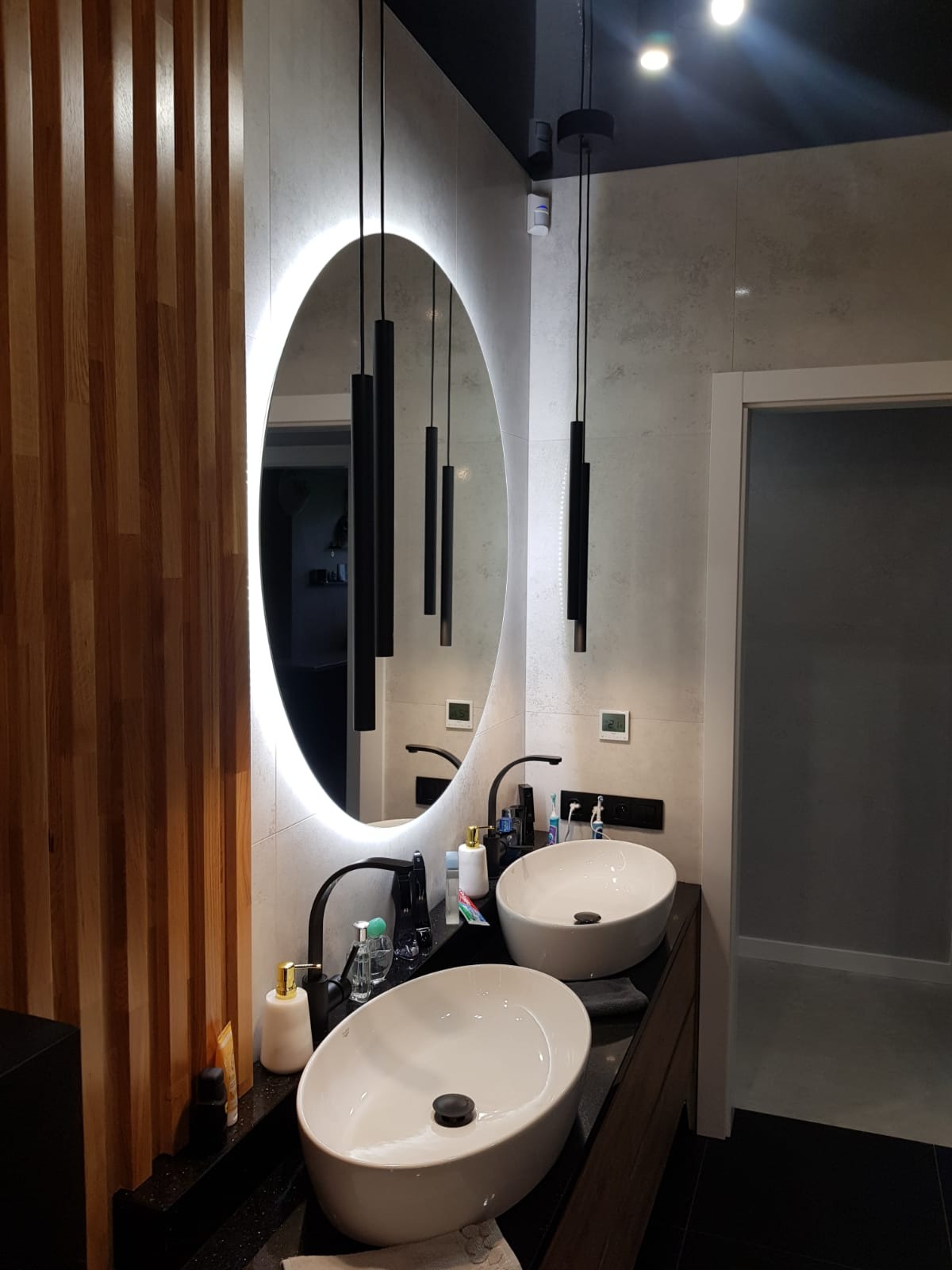 Nowoczesna łazienka z dwoma owalnymi umywalkami nablatowymi, czarnymi bateriami i podświetlanym owalnym lustrem. Ściana wykończona płytkami imitującymi kamień, a obok drewniane lamele.