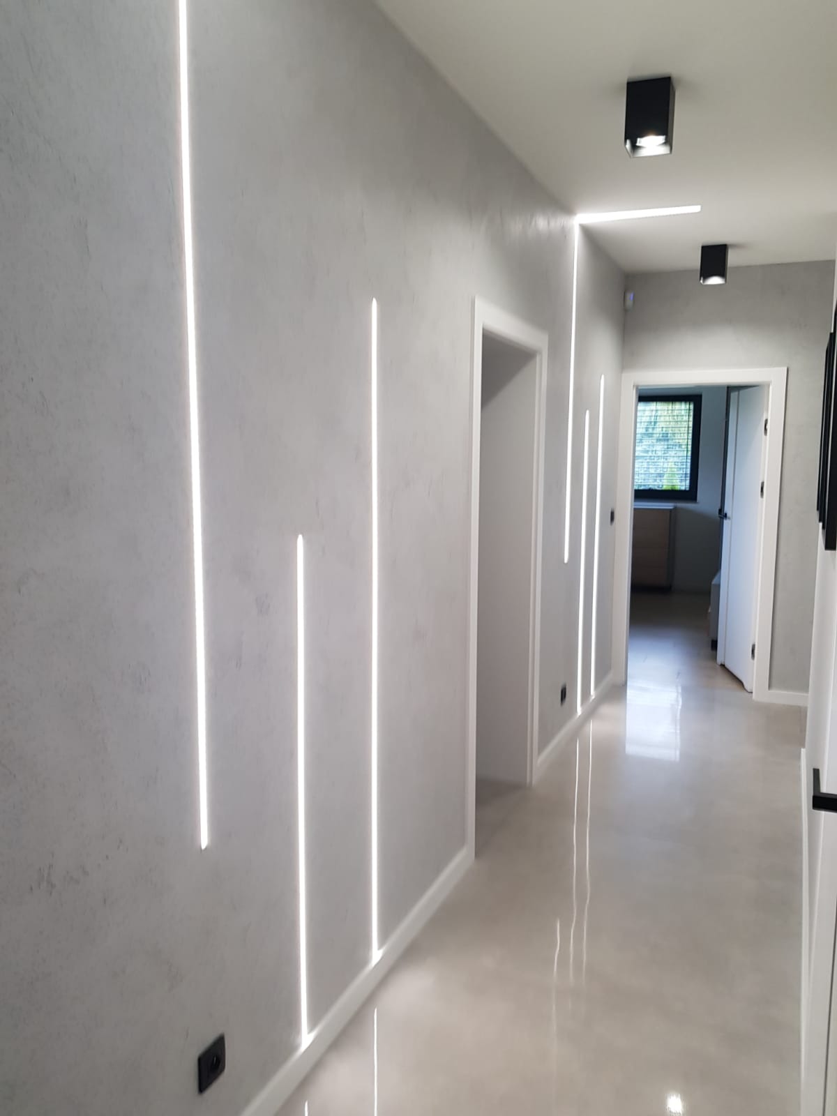 Korytarz z szarą ścianą o fakturze betonu, oświetlony pionowymi pasami LED, z widocznymi drzwiami i czarnymi lampami sufitowymi.