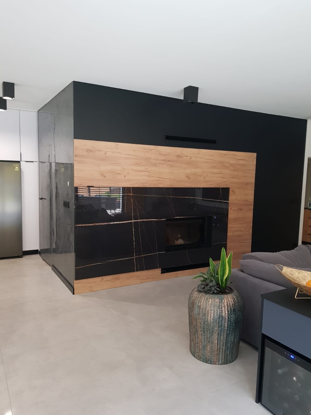 Nowoczesny salon z czarną ścianą, obudową kominka z forniru drewnopodobnego i czarnego marmuru z złotymi żyłami, szarą podłogą i rośliną doniczkową w ozdobnej osłonce.