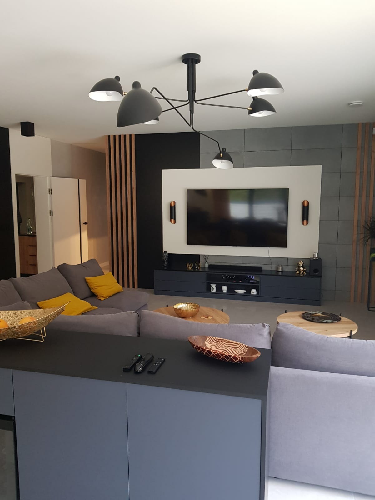 Nowoczesny salon z szarą sofą, drewnianymi lamelami dekoracyjnymi, czarną ścianą i designerską lampą sufitową z kilkoma kloszami.