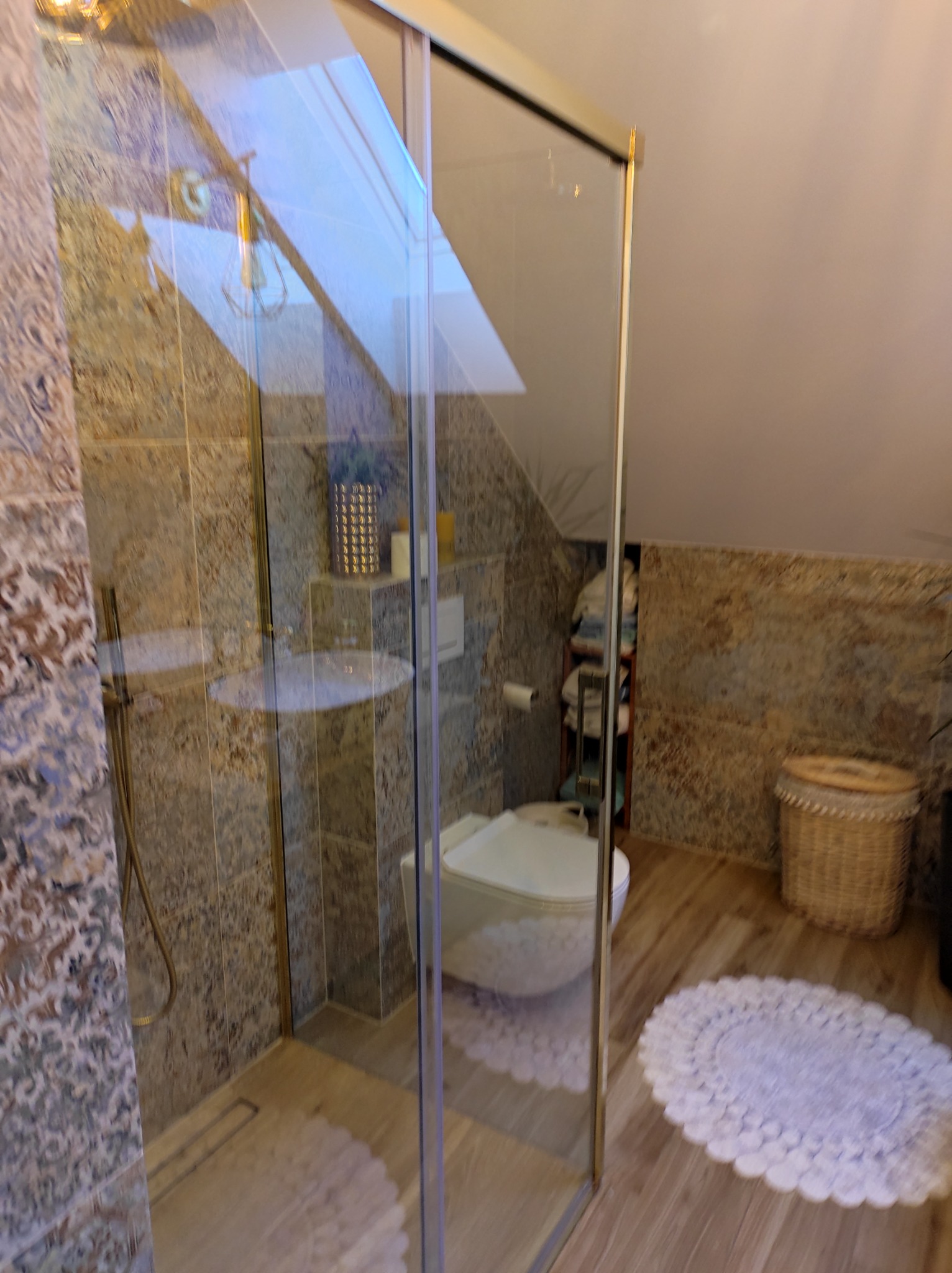 Nowoczesna łazienka na poddaszu z kabiną prysznicową ze złotymi profilami, toaletą typu Geberit, dekoracyjnymi płytkami ściennymi i drewnopodobną podłogą.