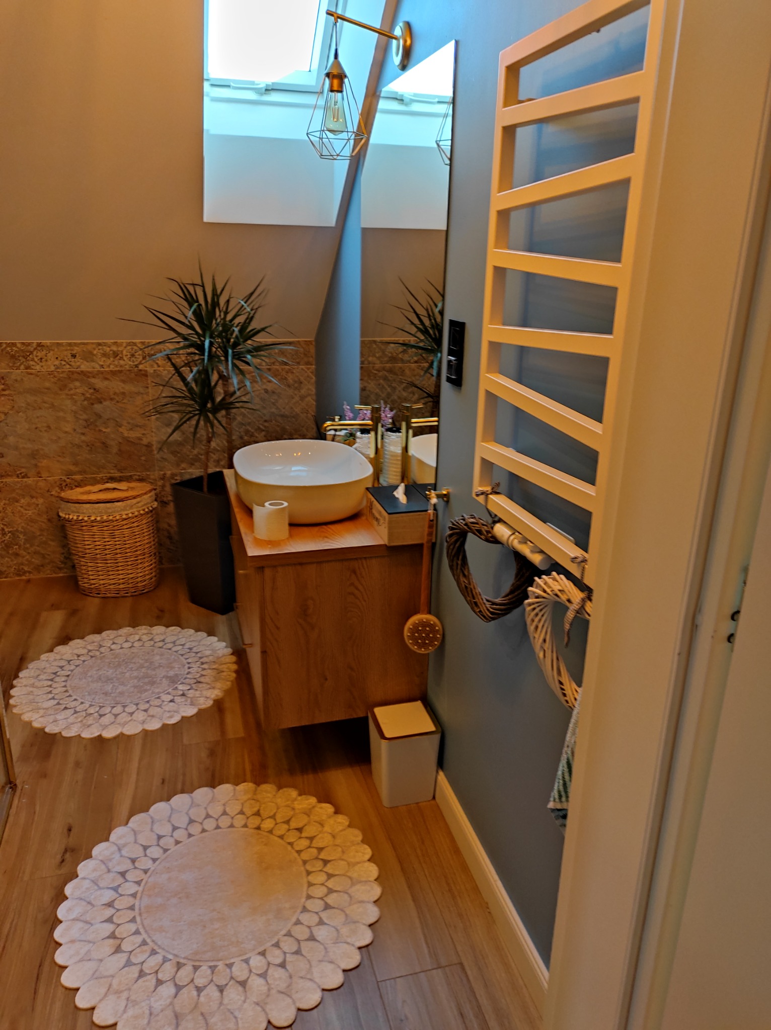 Jasna łazienka z drewnianą podłogą i okrągłymi dywanikami, biała umywalka nablatowa na szafce z jasnego drewna, grzejnik drabinkowy z dekoracjami, roślina w czarnej donicy, oraz lampa wisząca...