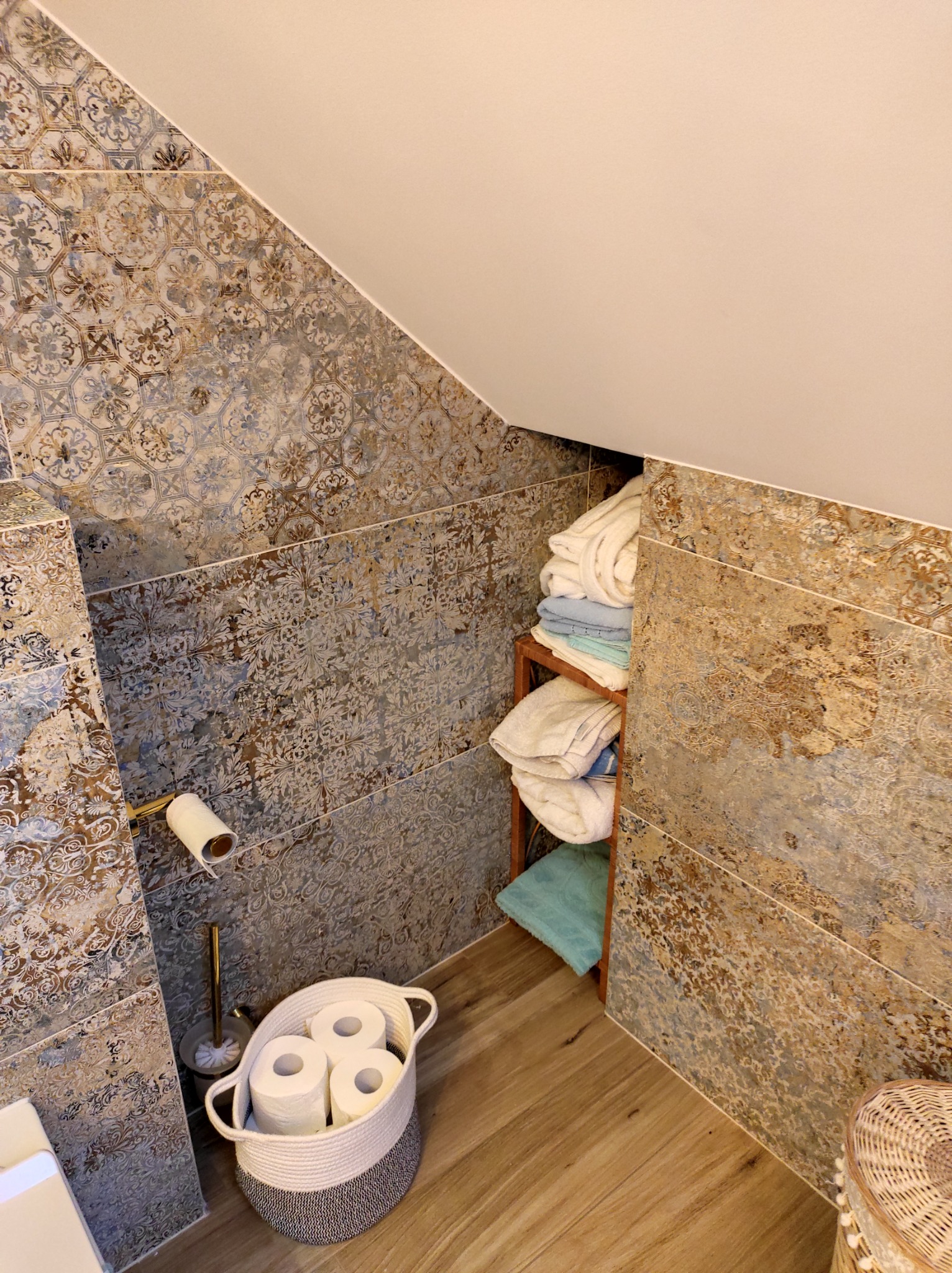 Ukośna ściana łazienki wyłożona dekoracyjnymi, wzorzystymi płytkami w odcieniach brązu i szarości, z drewnianą półką z ręcznikami i koszem z zapasem papieru toaletowego.
