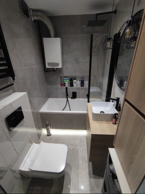 Nowoczesna łazienka z szarymi płytkami, białą wanną z czarną baterią, umywalką nablatową i podświetleniem LED pod wanną, wisząca toaleta z czarnym przyciskiem oraz dekoracyjne lampy sufitowe.