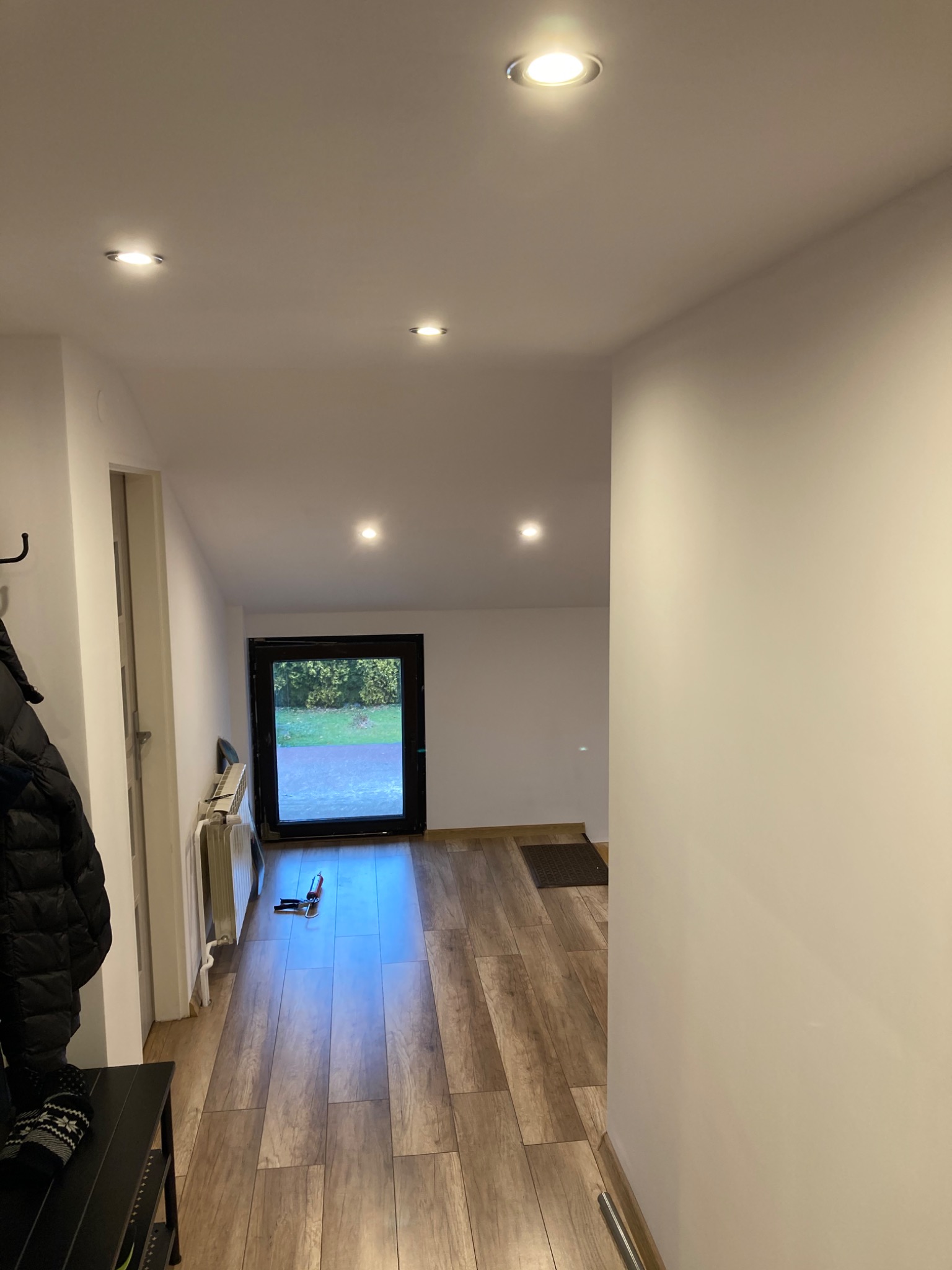 Korytarz po remoncie z panelami podłogowymi imitującymi drewno, białymi ścianami, czarną ramą drzwi wejściowych i wbudowanymi lampami sufitowymi; na podłodze leżą narzędzia.