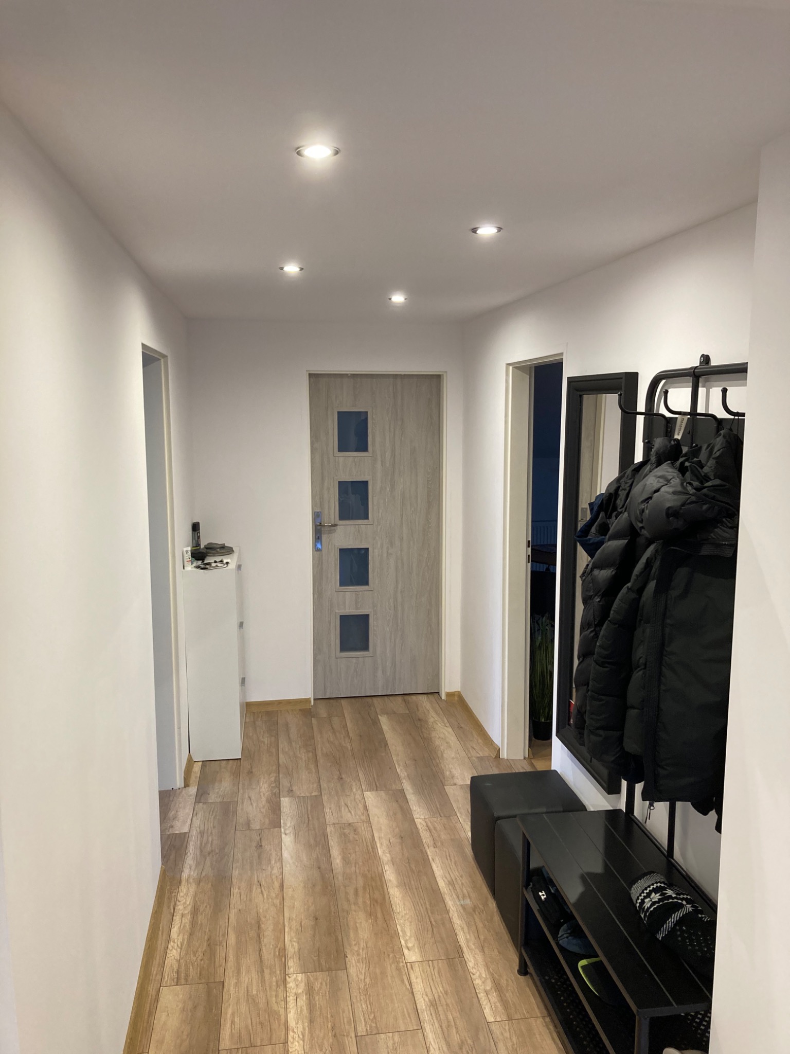 Nowoczesny przedpokój z panelami podłogowymi imitującymi drewno, białe ściany, drzwi wejściowe z czterema szybkami, czarny wieszak na ubrania z lustrem i pufą.