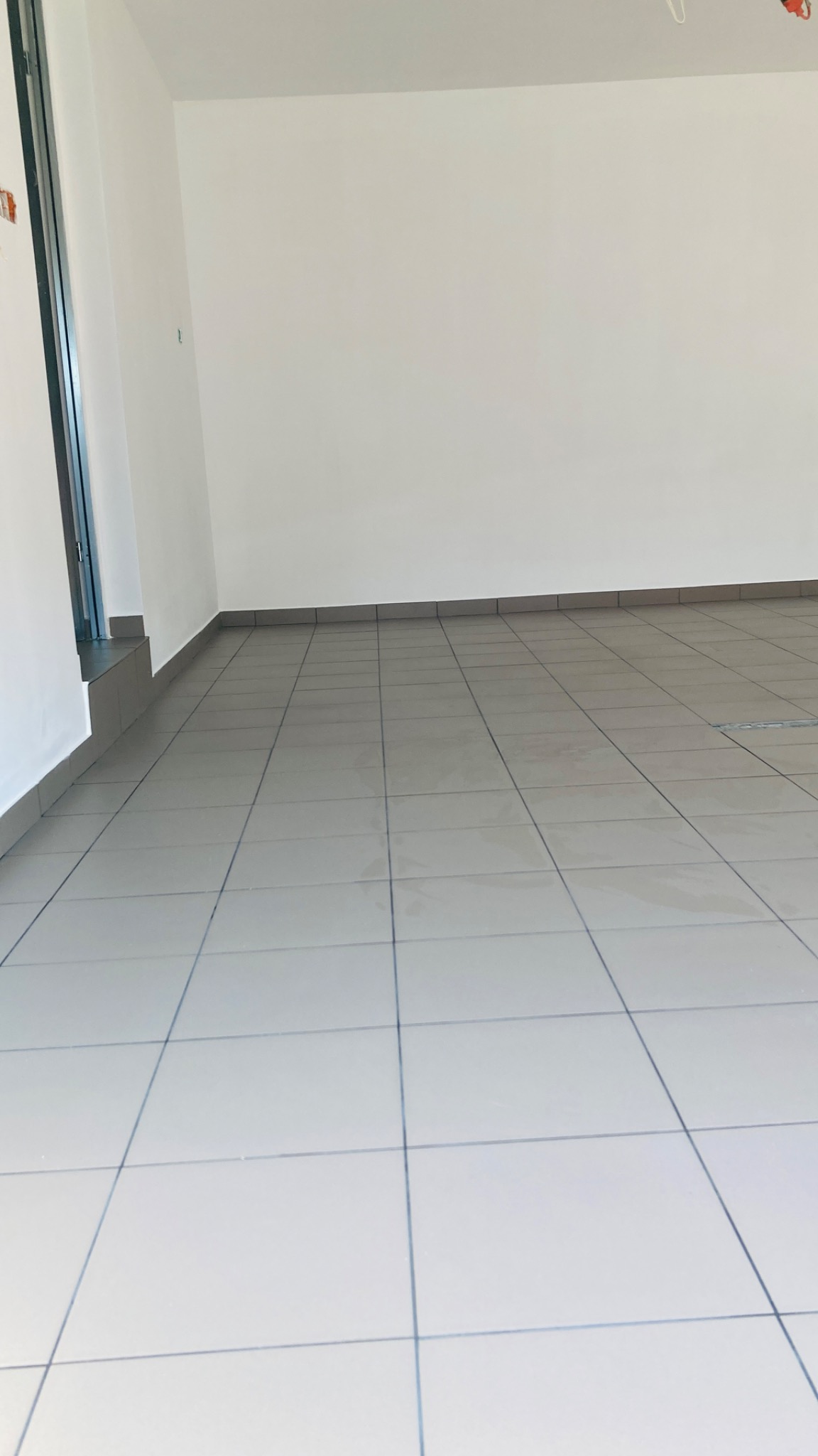 Jasnoszare, błyszczące płytki podłogowe ułożone w kwadraty, z ciemnoszarą listwą przypodłogową w pustym, odnowionym pomieszczeniu z białymi ścianami.