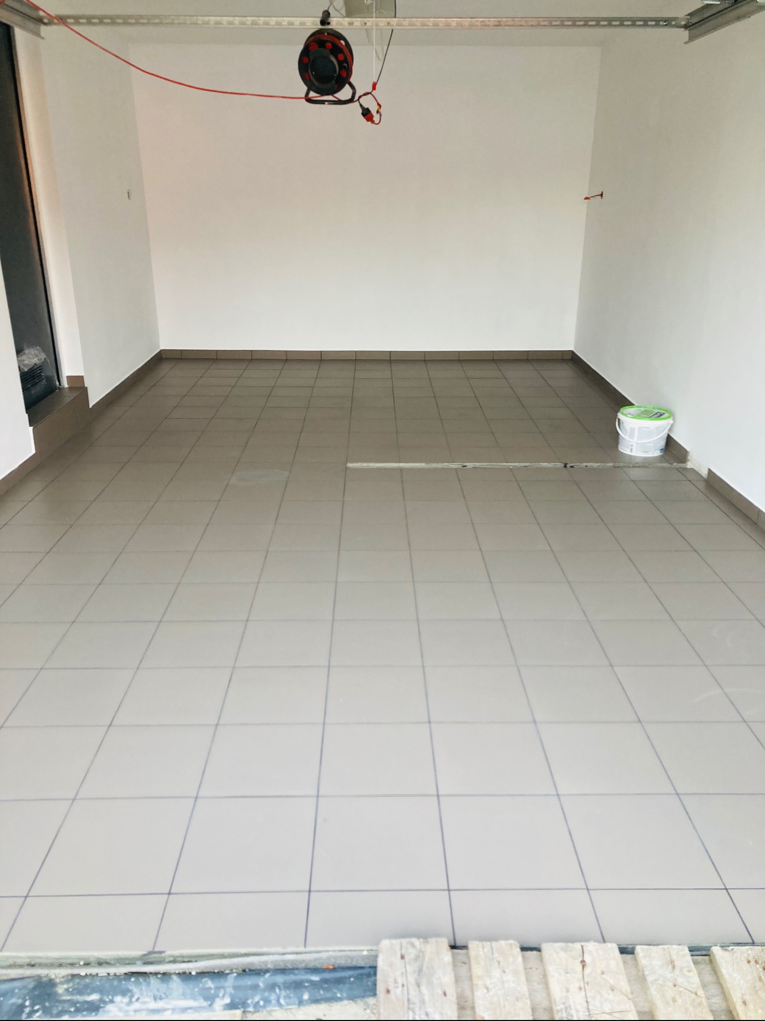 Wyłożona szarymi płytkami podłoga w pustym pomieszczeniu, widoczny bęben z kablem elektrycznym pod sufitem i wiadro z farbą przy ścianie.