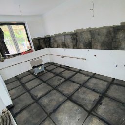 WJ Waszka - Pomieszczenie w trakcie remontu z ułożonymi ciemnymi płytkami podłogowymi i ściennymi, widocznymi miedzianymi rurami instalacji wodnej oraz oknem częściowo zakrytym folią.