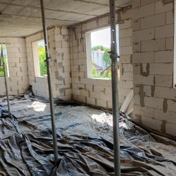 WJ Waszka - Wnętrze budowanego domu z białych bloczków, z betonowym stropem podpartym trzema metalowymi stemplami. Na podłodze pofałdowana czarna folia, przez okna widać zieleń.