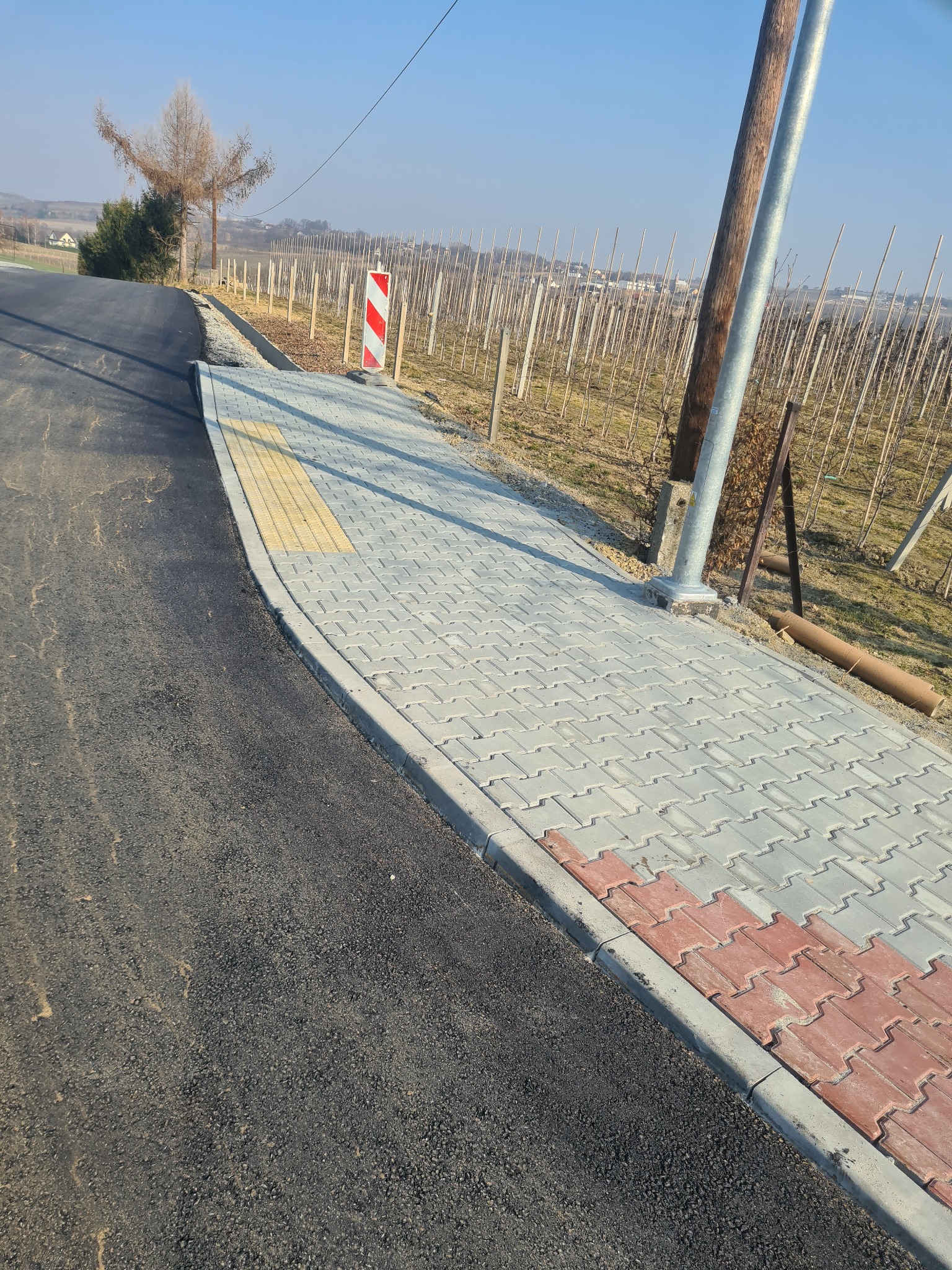 Świeżo ułożona kostka brukowa w Łącku, przylegająca do asfaltowej drogi, z elementami dla osób niedowidzących i słupem oświetleniowym, w tle widok na sad.