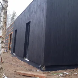 Ciemna, drewniana elewacja budynku w trakcie budowy, widoczne okna i fragment ocieplenia, teren wokół w stanie surowym.