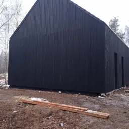 Czarny, nowoczesny budynek z drewnianą elewacją w trakcie budowy, widoczne deski na ziemi i surowy teren.